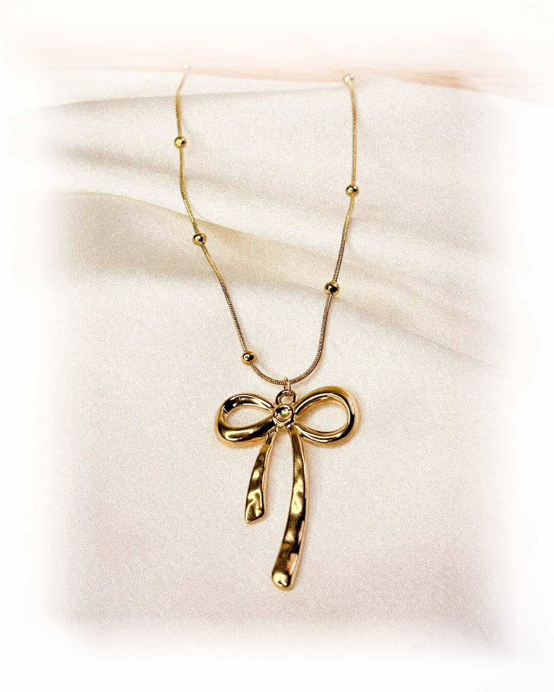 Elegant gold bow pendant necklace with beaded chain detail displayed on a soft white fabric background