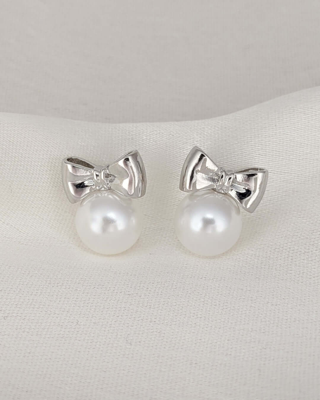Minimal silver bow earrings featuring pearl accents on a luxury white backdrop