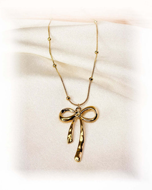 Elegant gold bow pendant necklace with beaded chain detail displayed on a soft white fabric background