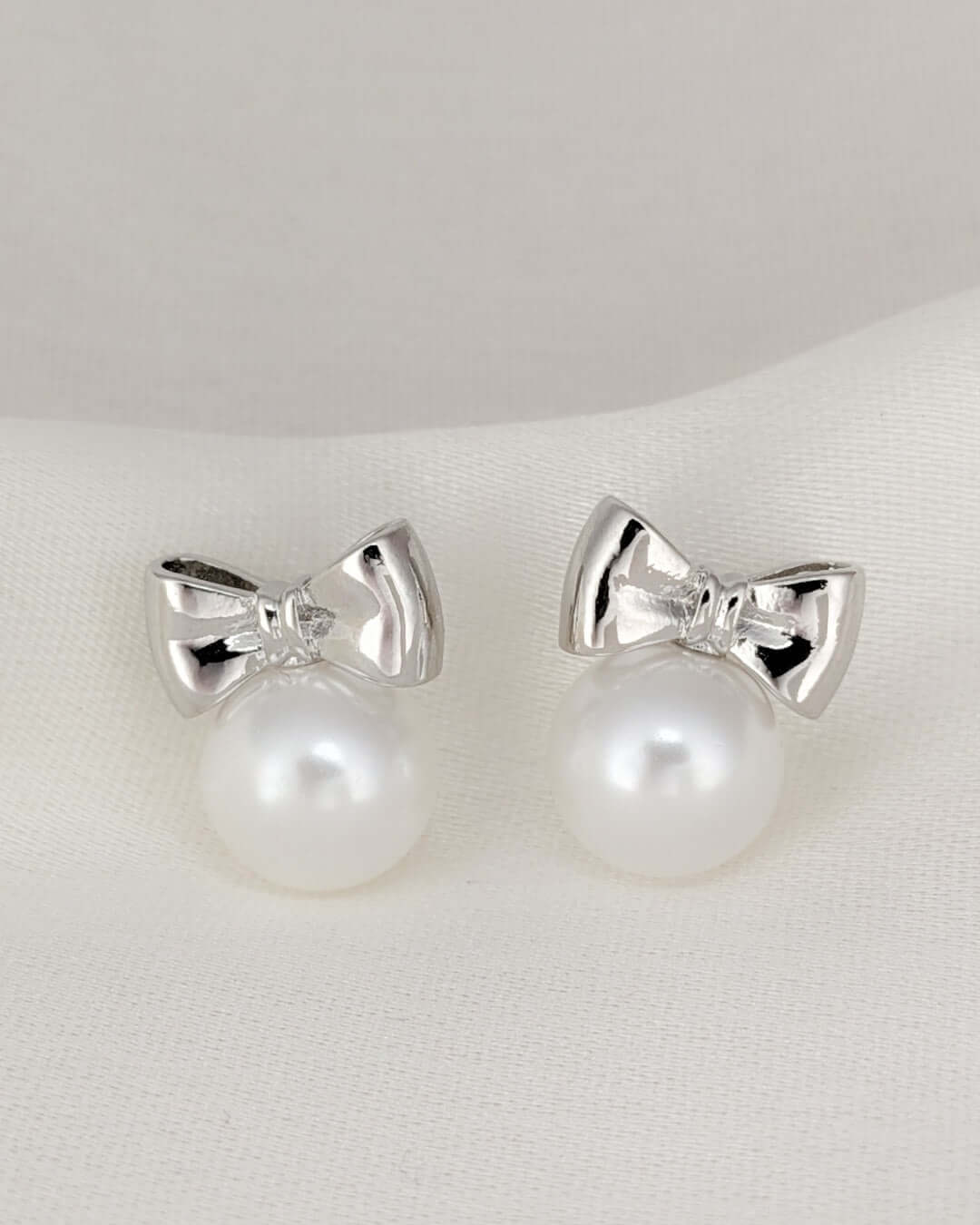 Silver bow stud earrings with pearl drops displayed on a soft white fabric background