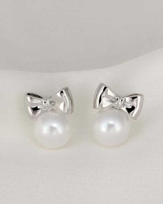 Silver bow stud earrings with pearl drops displayed on a soft white fabric background